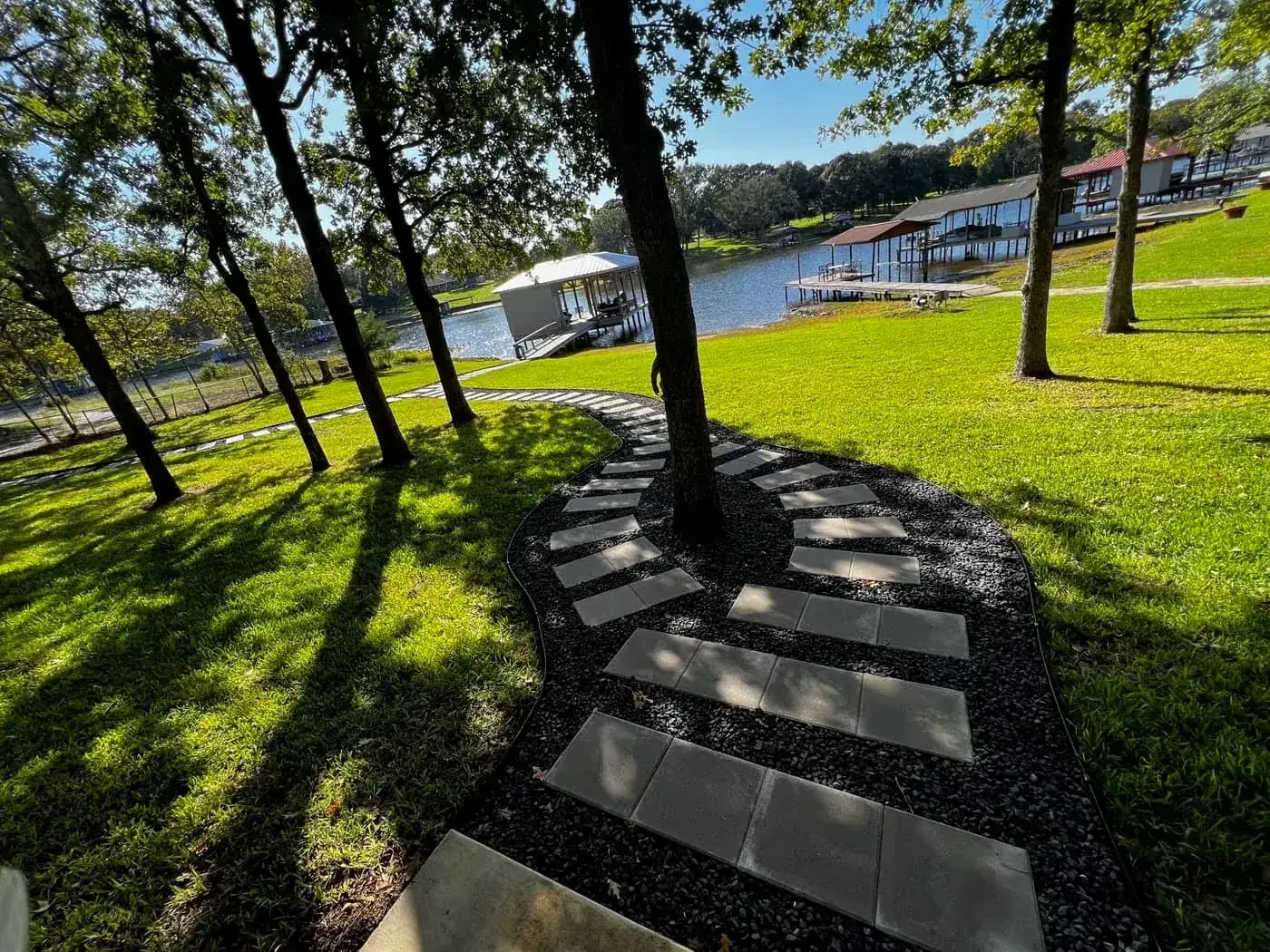Curved stepping-stone walkway through black river rock and lawn beneath oak trees, leading to a boathouse on the lake